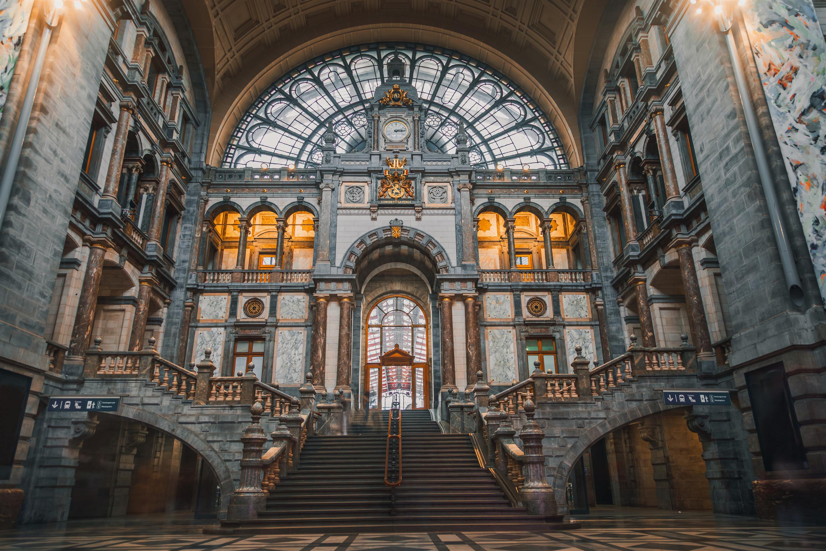 The concourse of a monumental railway terminus, soaring glass and ironwork filled with daylight, travellers small beneath it.