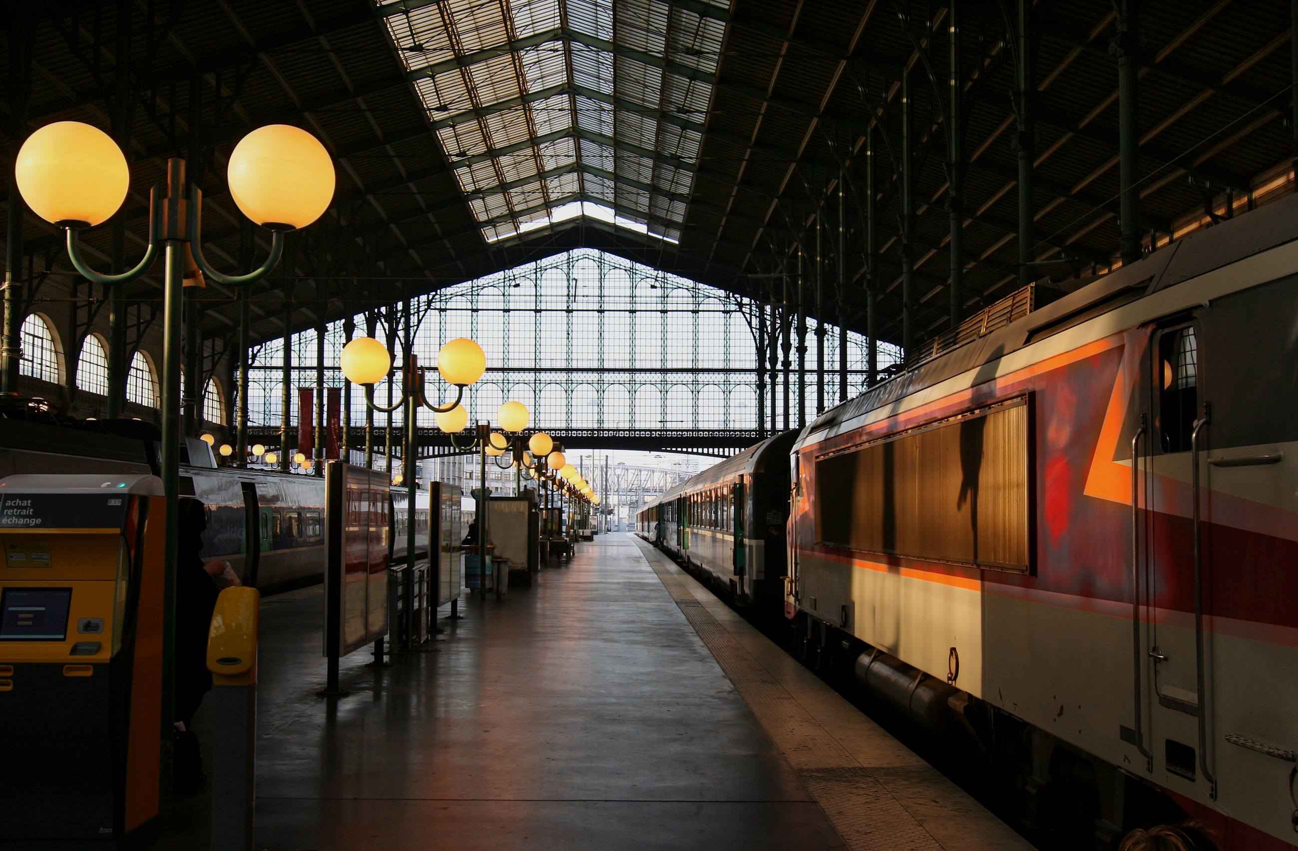A station platform under an arched glass roof, lamps receding into the distance and a train waiting alongside.