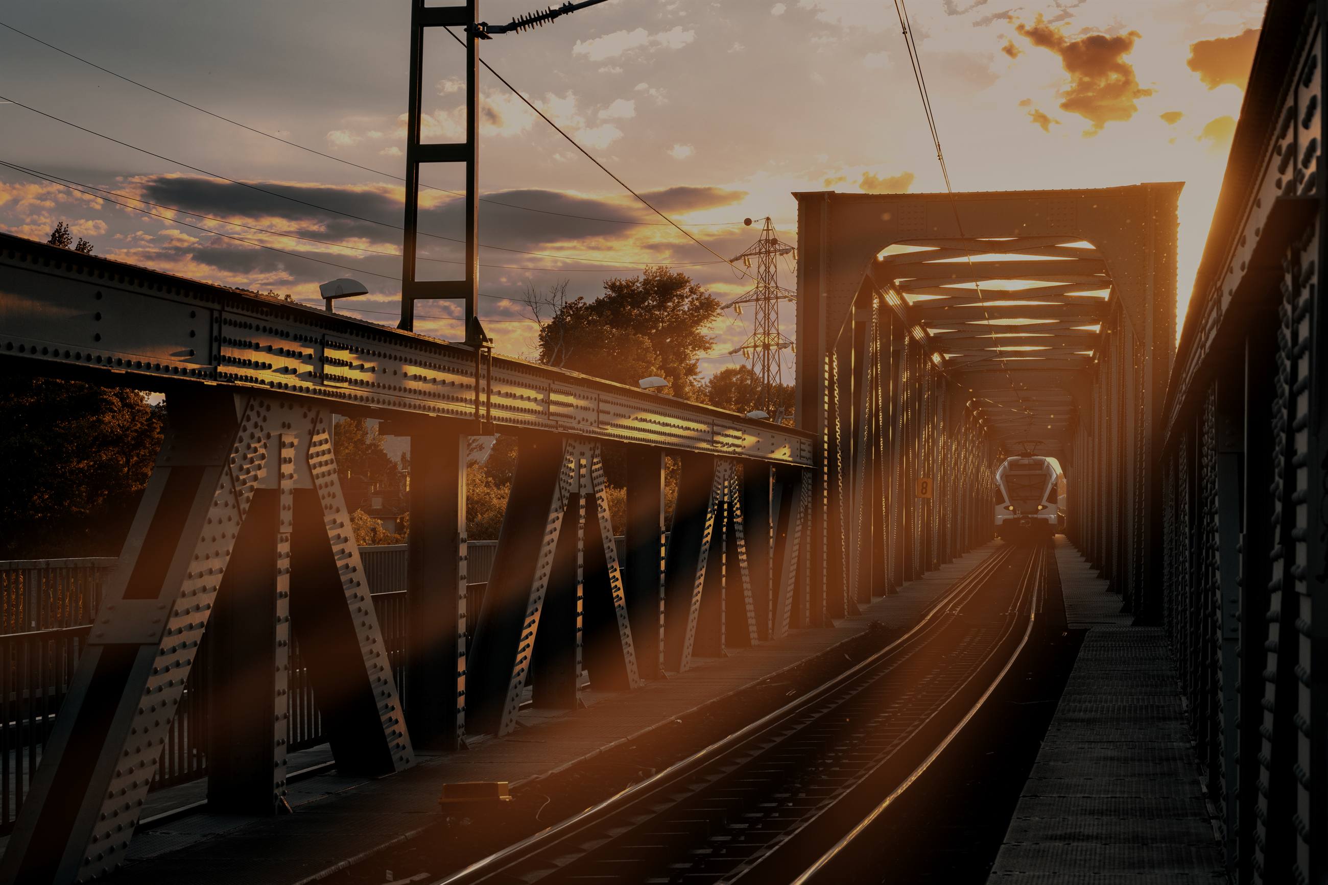 A steel railway bridge at sunset, a train approaching through the girders.