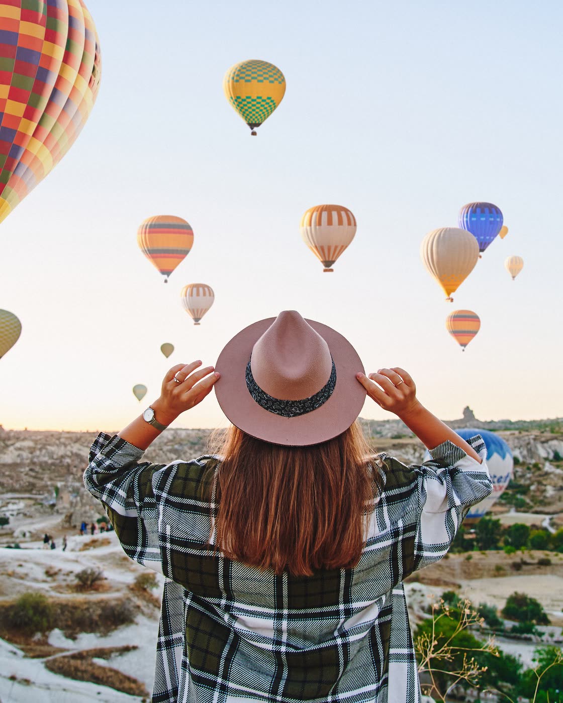 A woman in a hat watches a sky full of hot air balloons at sunrise.
