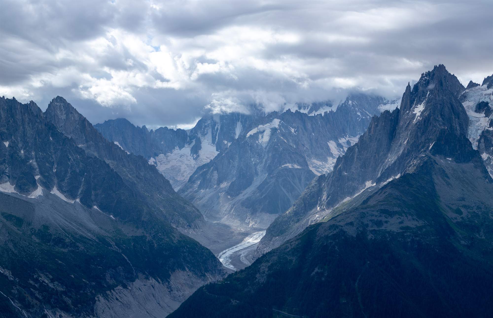 A glacier running down a valley between two dark mountain masses, cloud closing over the peaks above.