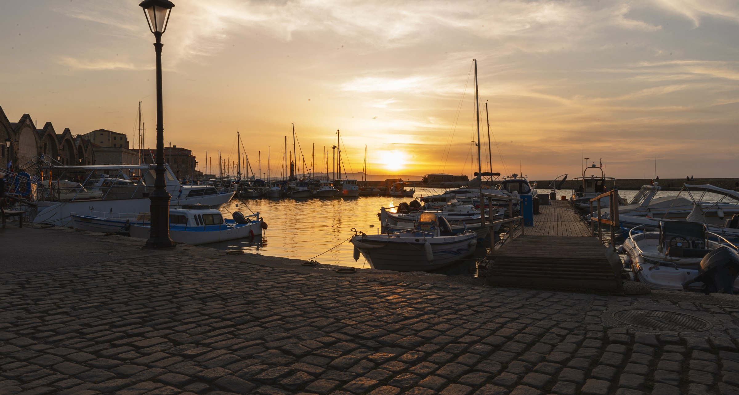 A stone harbour quay at sunset, a lamp post lit against the water and boats moored along the dock.