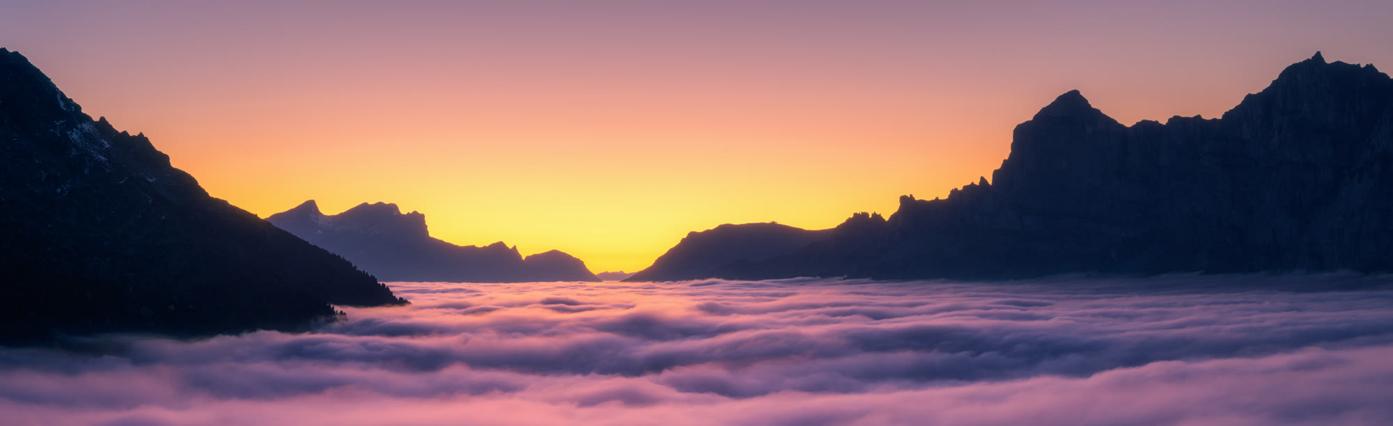 A sea of cloud filling an alpine valley between dark peaks, the sky gold at the horizon before the sun has risen.