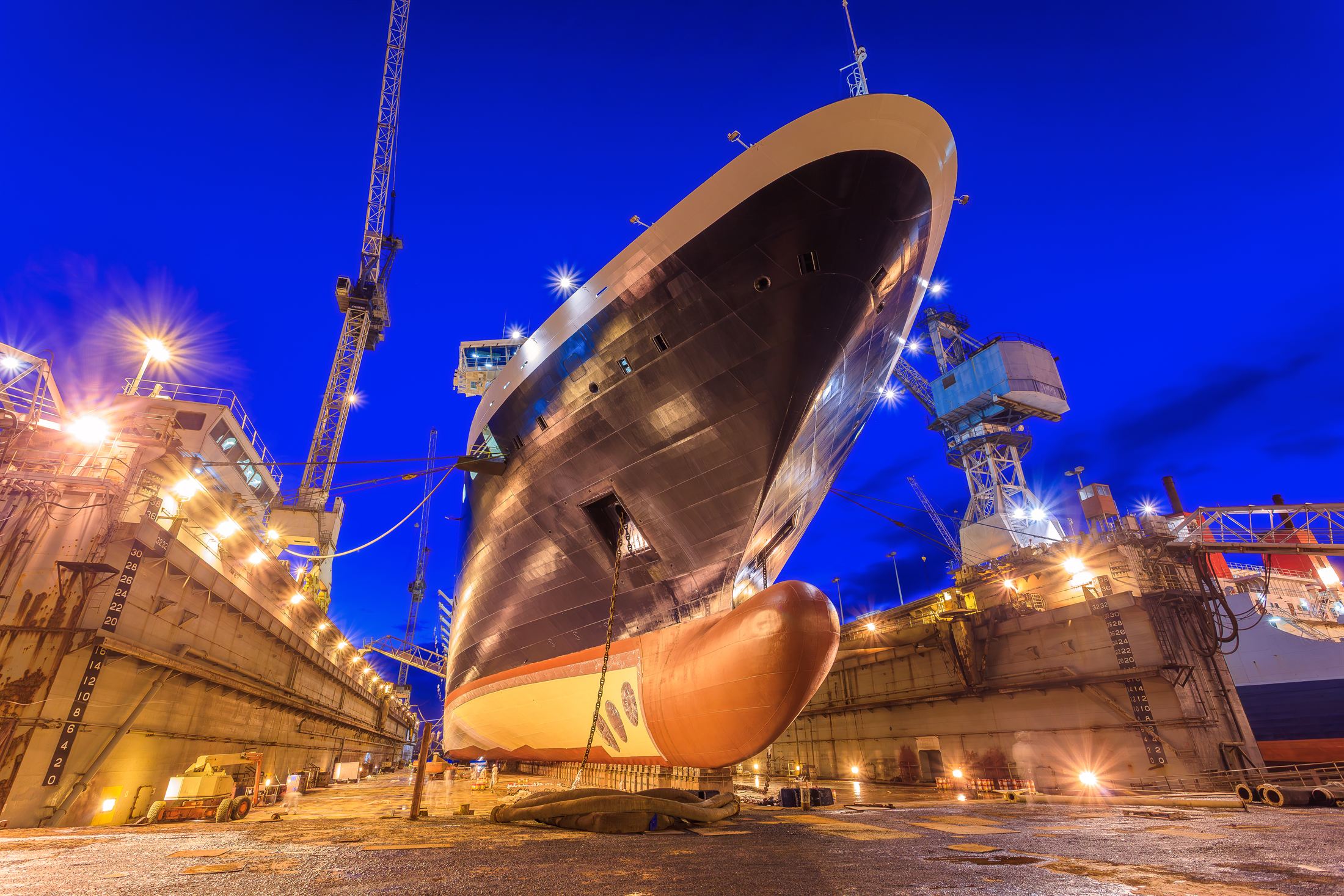 A ship raised in dry dock at blue hour, cranes and work lights burning around its hull.