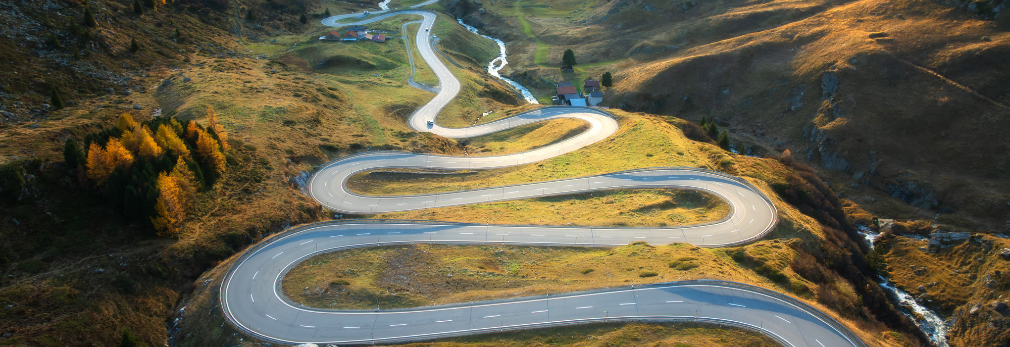 A mountain pass road looping up an autumn hillside in low sun, seen from above, the valley floor and a farm beyond it.
