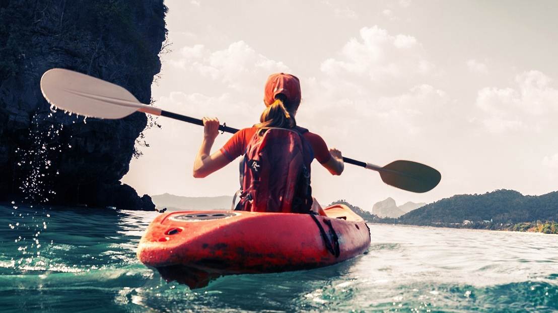 A kayaker paddles alone through calm turquoise water beneath towering cliffs.