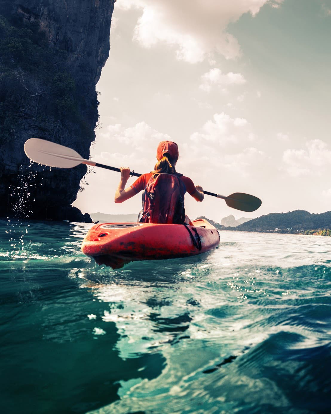 A kayaker paddles alone through calm turquoise water beneath towering cliffs.