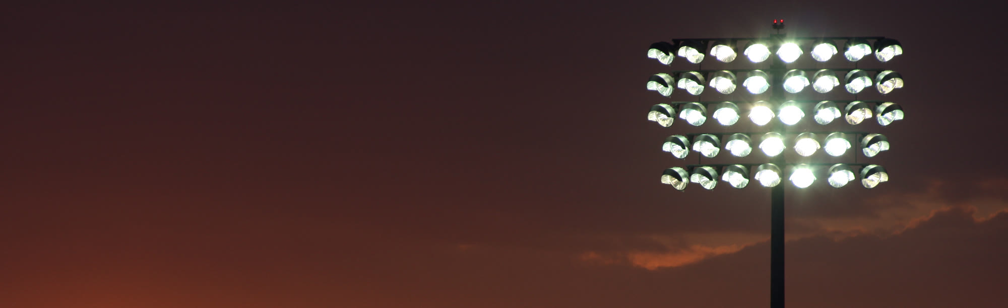 A single floodlight rig lit against an amber sunset sky, a band of dark cloud behind it.