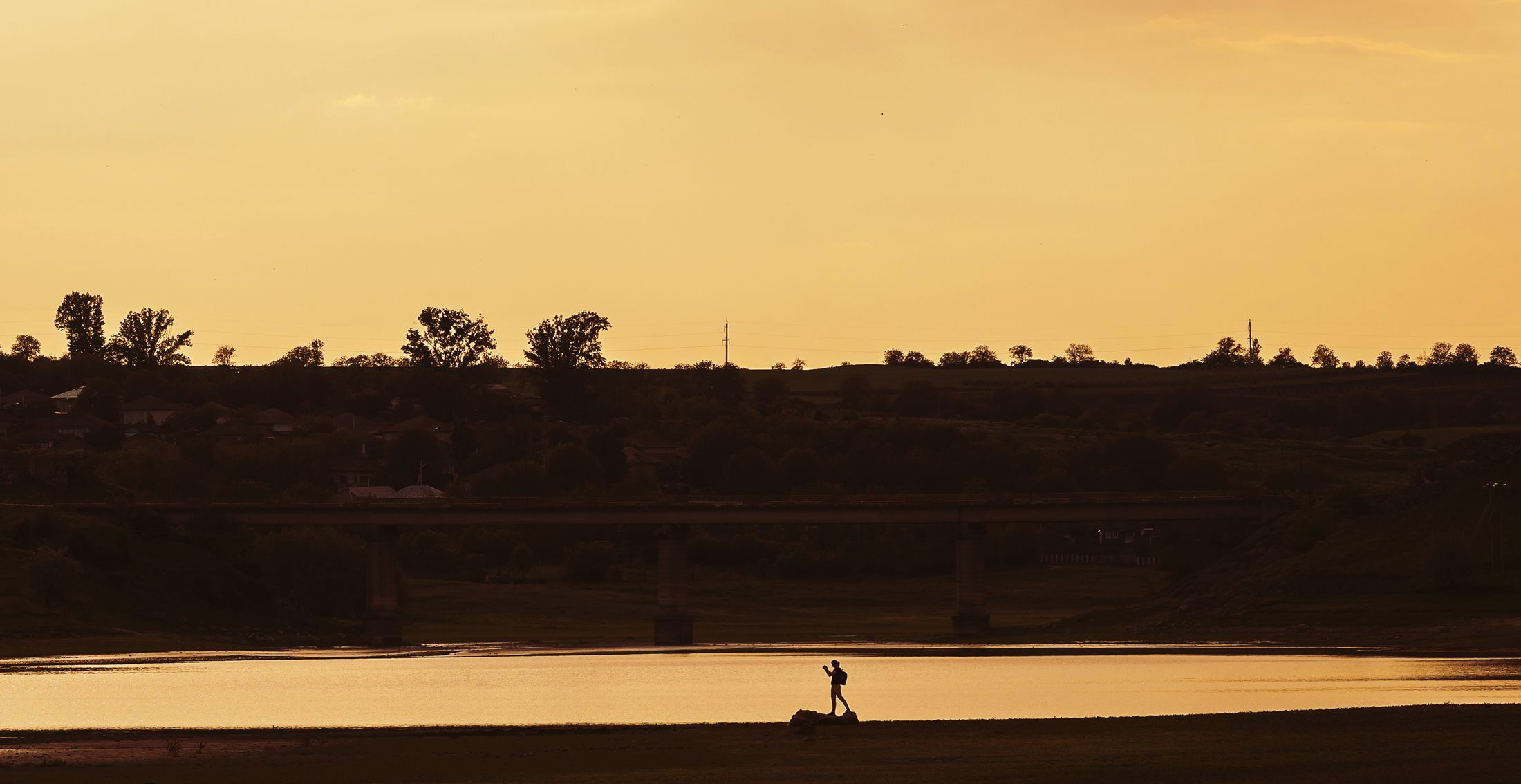 A single figure standing at the water's edge, alone against a vast bronze sky at sunset.