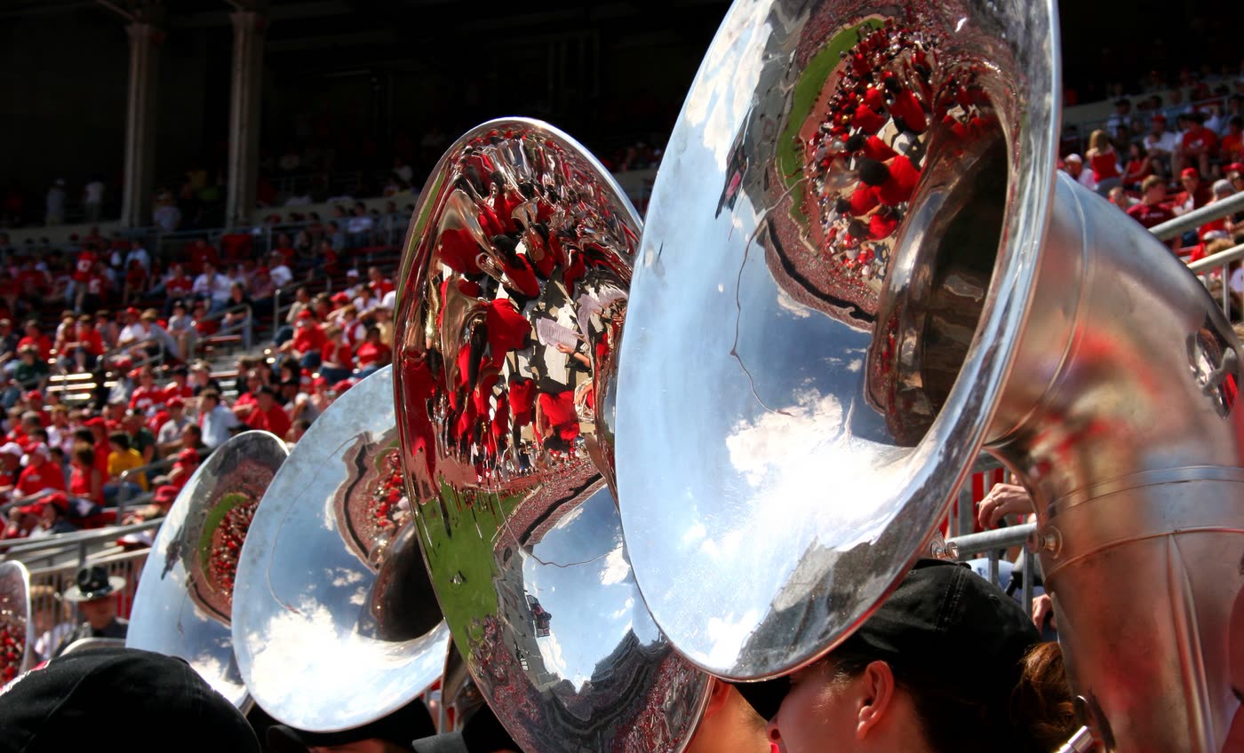 Sunlight and a crowd reflected in the polished bells of marching band tubas.
