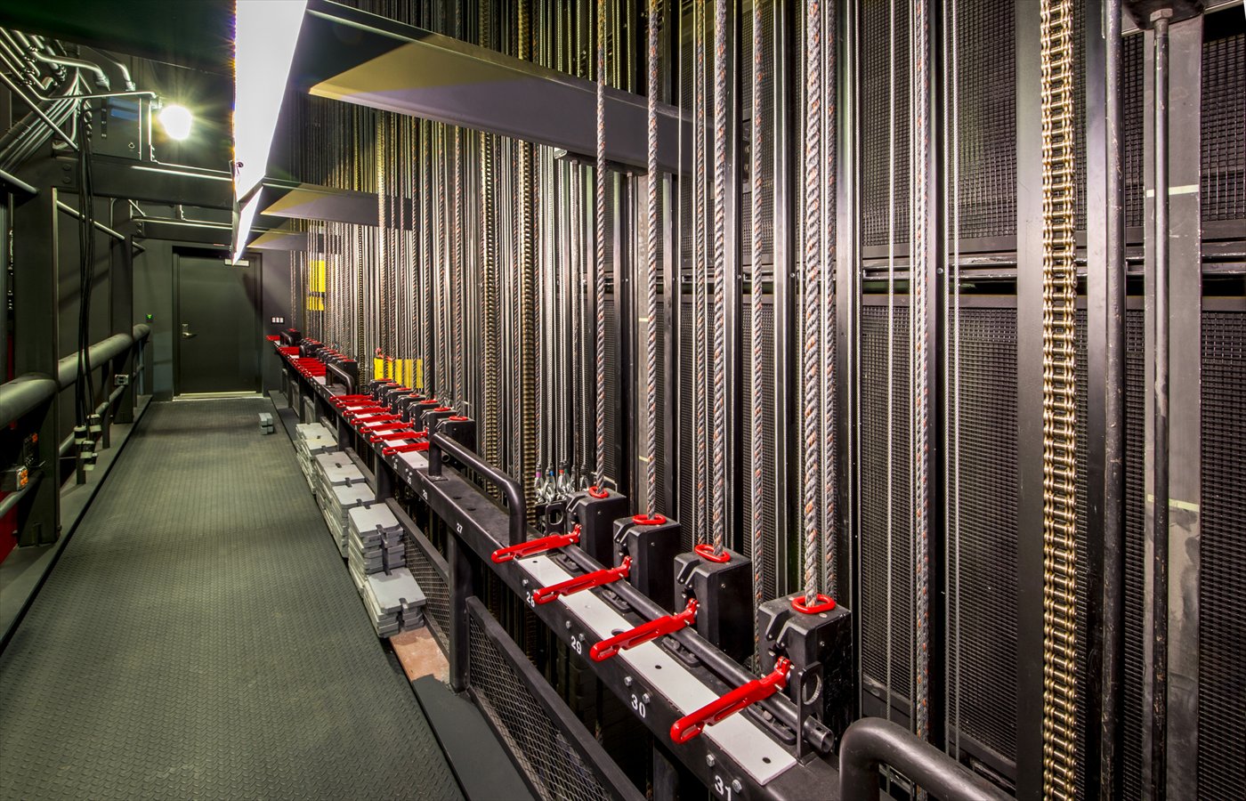A fly-system counterweight corridor, ropes and locking rails with red safety locks running the length of it.