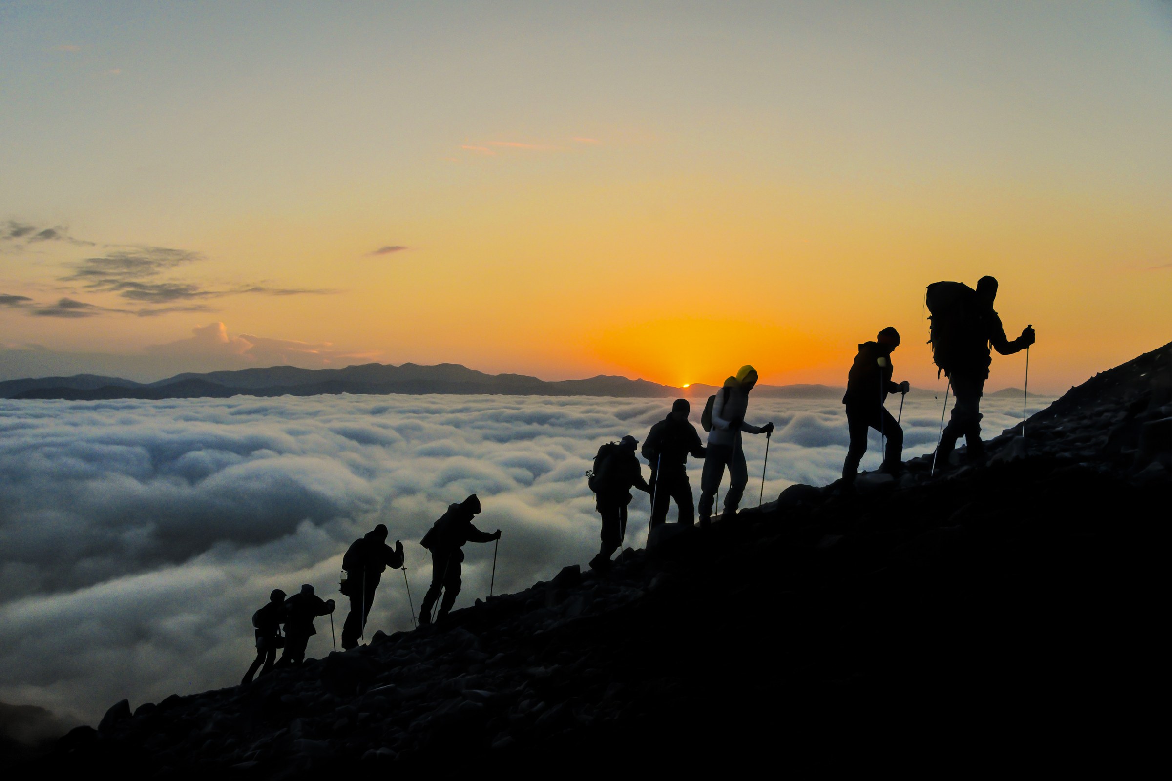 A line of hikers ascending a ridge above the clouds at sunset.