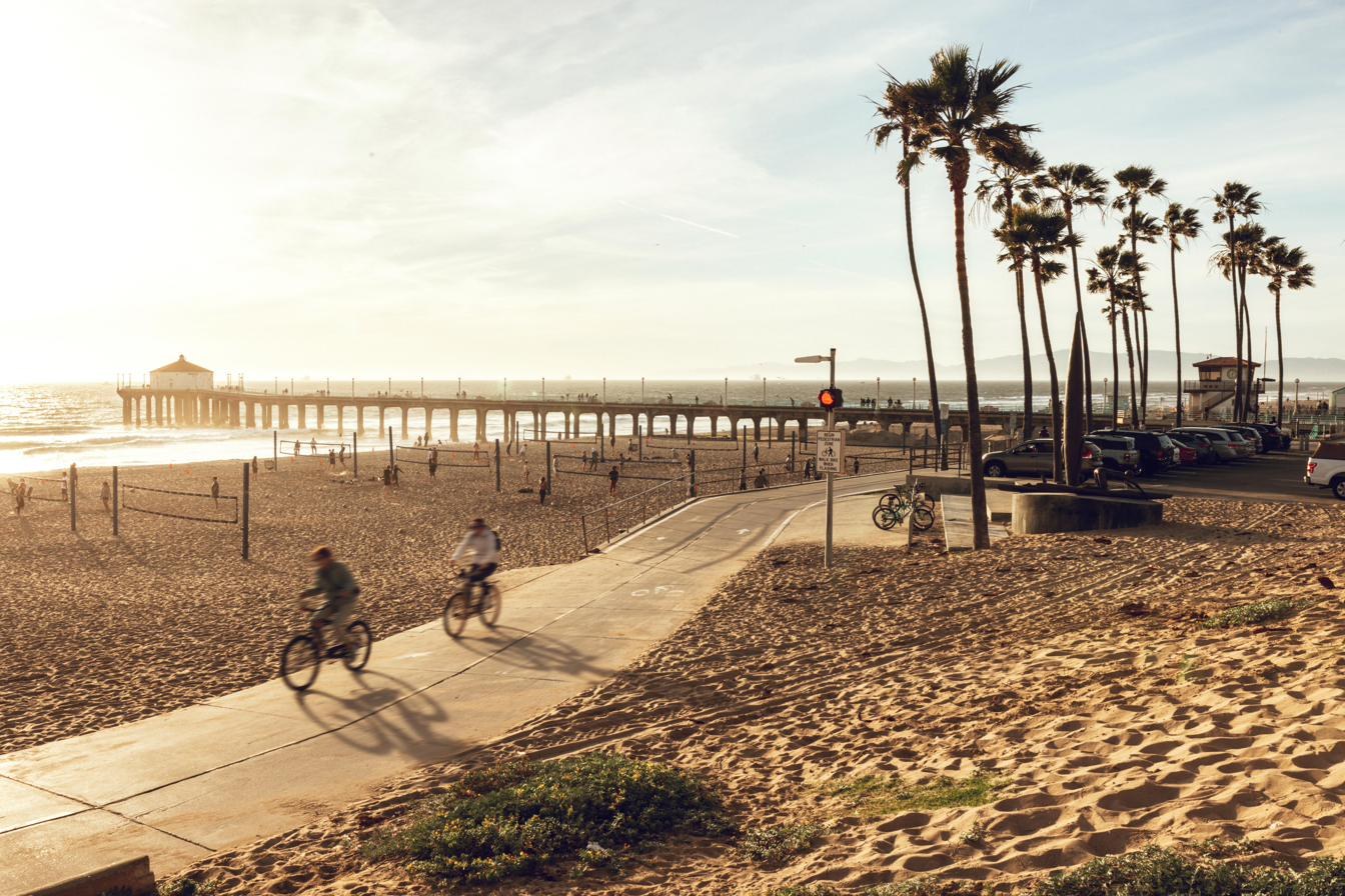 Cyclists ride the boardwalk past Manhattan Beach Pier at golden hour, palm trees lining the path.