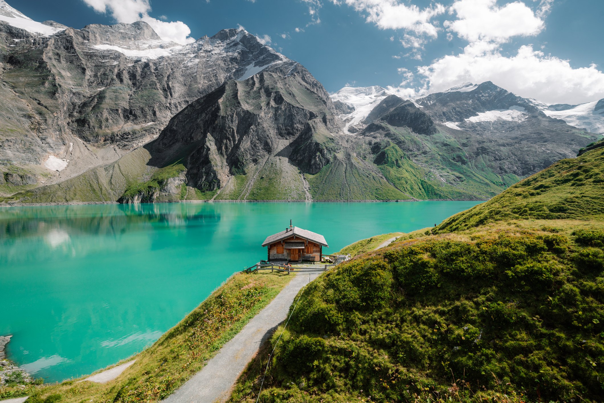 A turquoise glacial lake beneath snow-veined peaks, with a single wooden cabin on the shore and a footpath leading to it.
